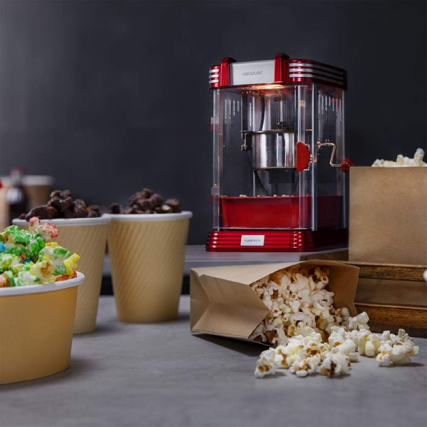 Macchina per popcorn rossa con sacchetti di carta e bicchieri di popcorn colorati su un tavolo grigio.