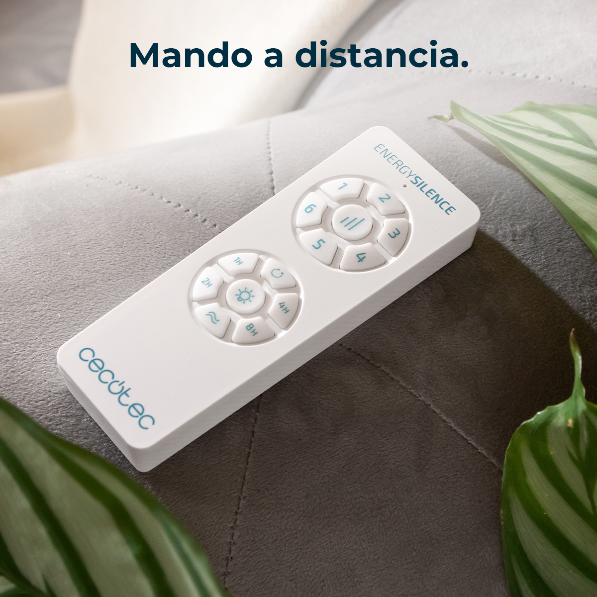 Un telecomando bianco Cecotec EnergySilence si trova su una superficie grigia tra foglie verdi.