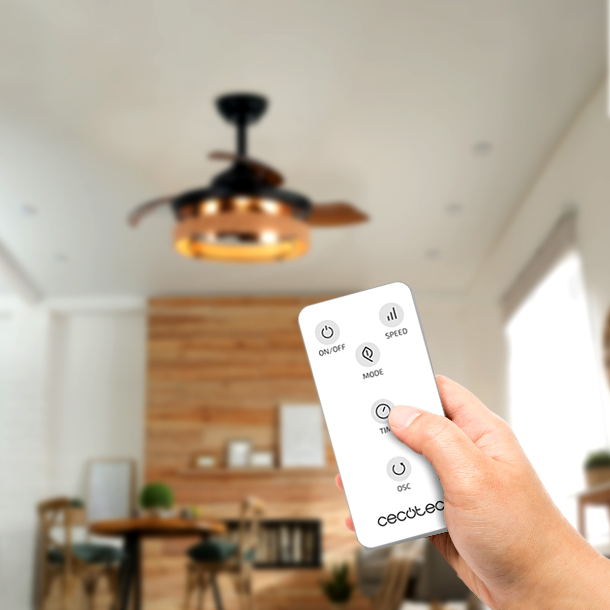 Una mano tiene un telecomando bianco puntato verso un ventilatore da soffitto illuminato con pale in legno.