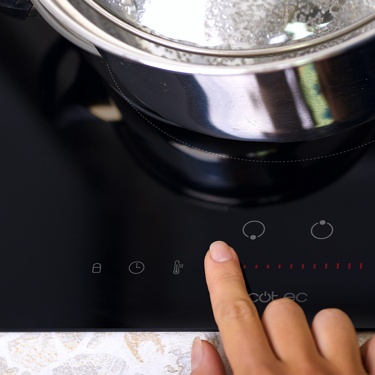 Una mano toca los controles táctiles de una moderna placa de inducción negra con una olla encima.
