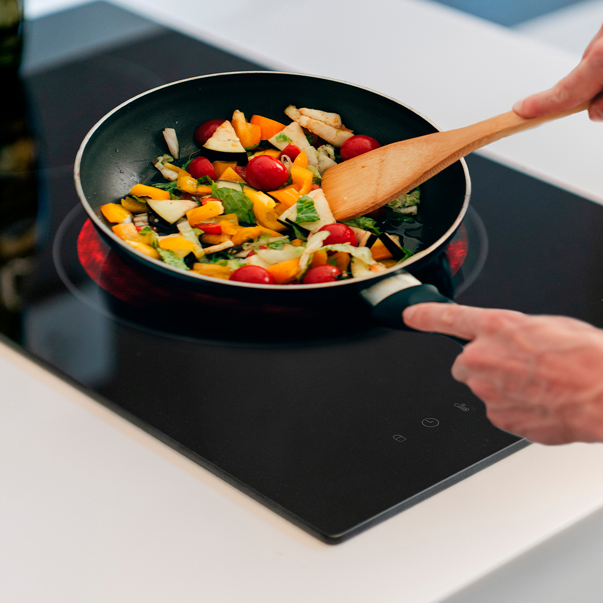 Una persona revuelve verduras coloridas con una espátula de madera en una sartén sobre una placa de inducción.