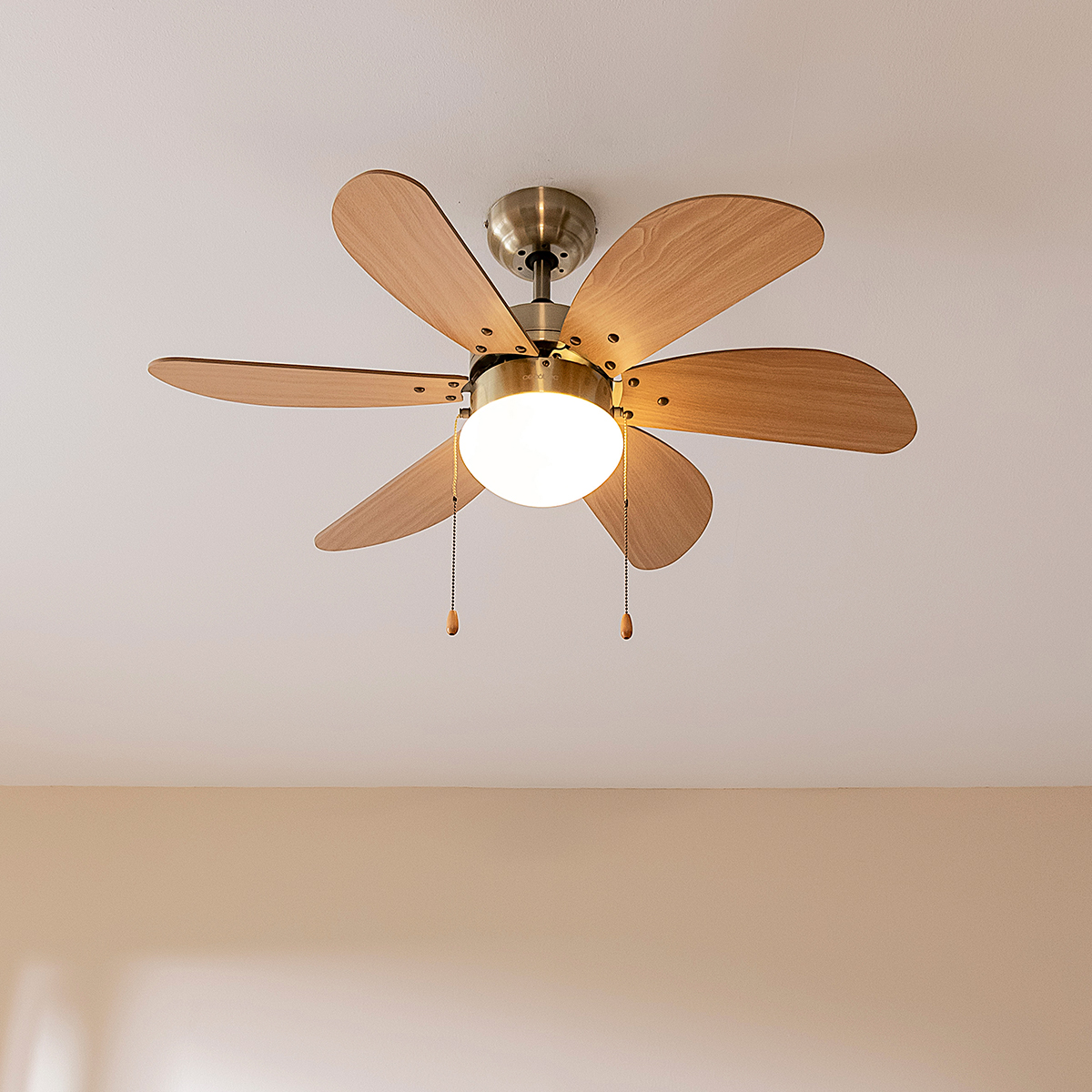 Un ventilatore da soffitto con sei pale in legno chiaro e una lampada rotonda integrata pende da un soffitto bianco.