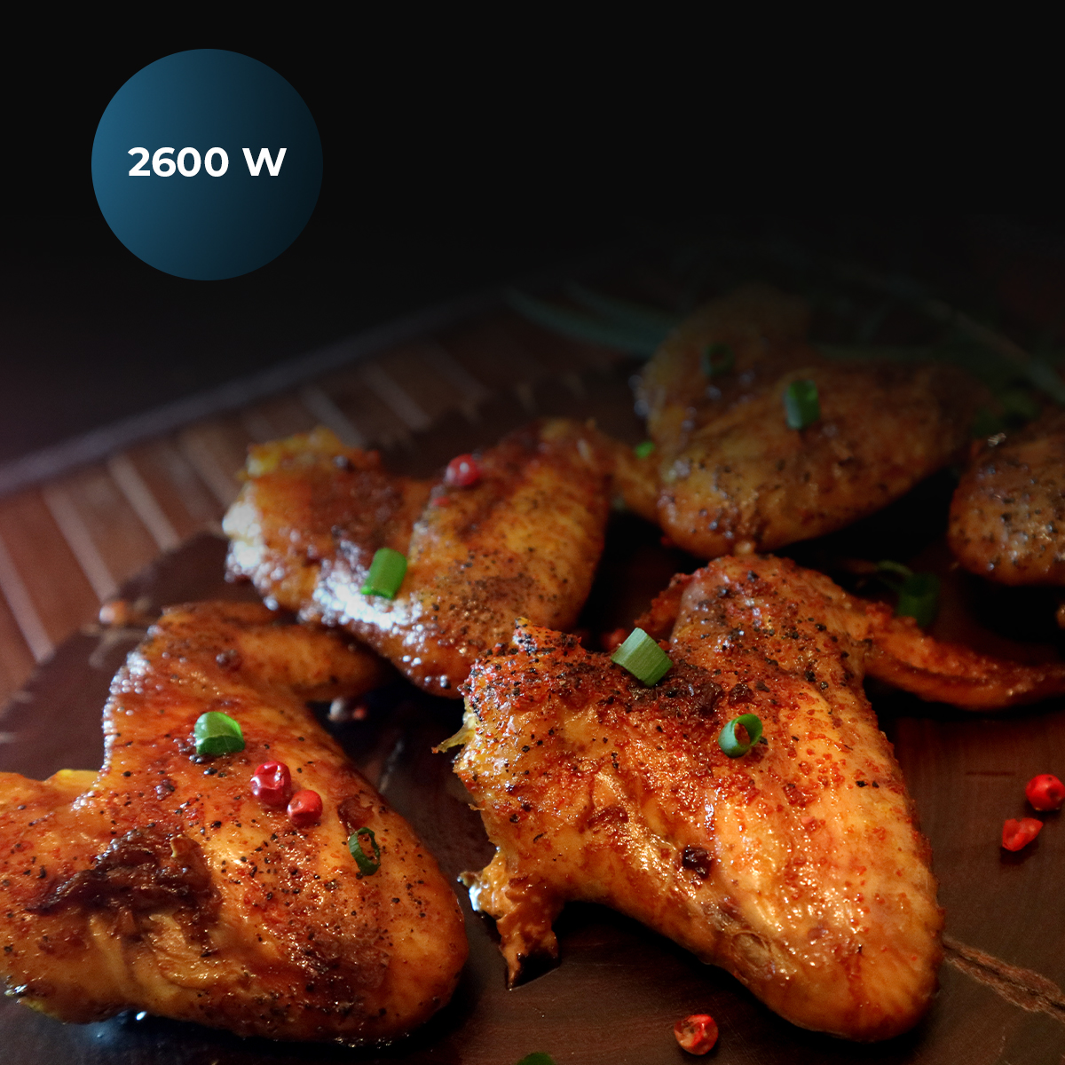 Alitas de pollo sazonadas en una tabla de madera, adornadas con cebollino y pimienta. Una insignia azul muestra 2600 W.
