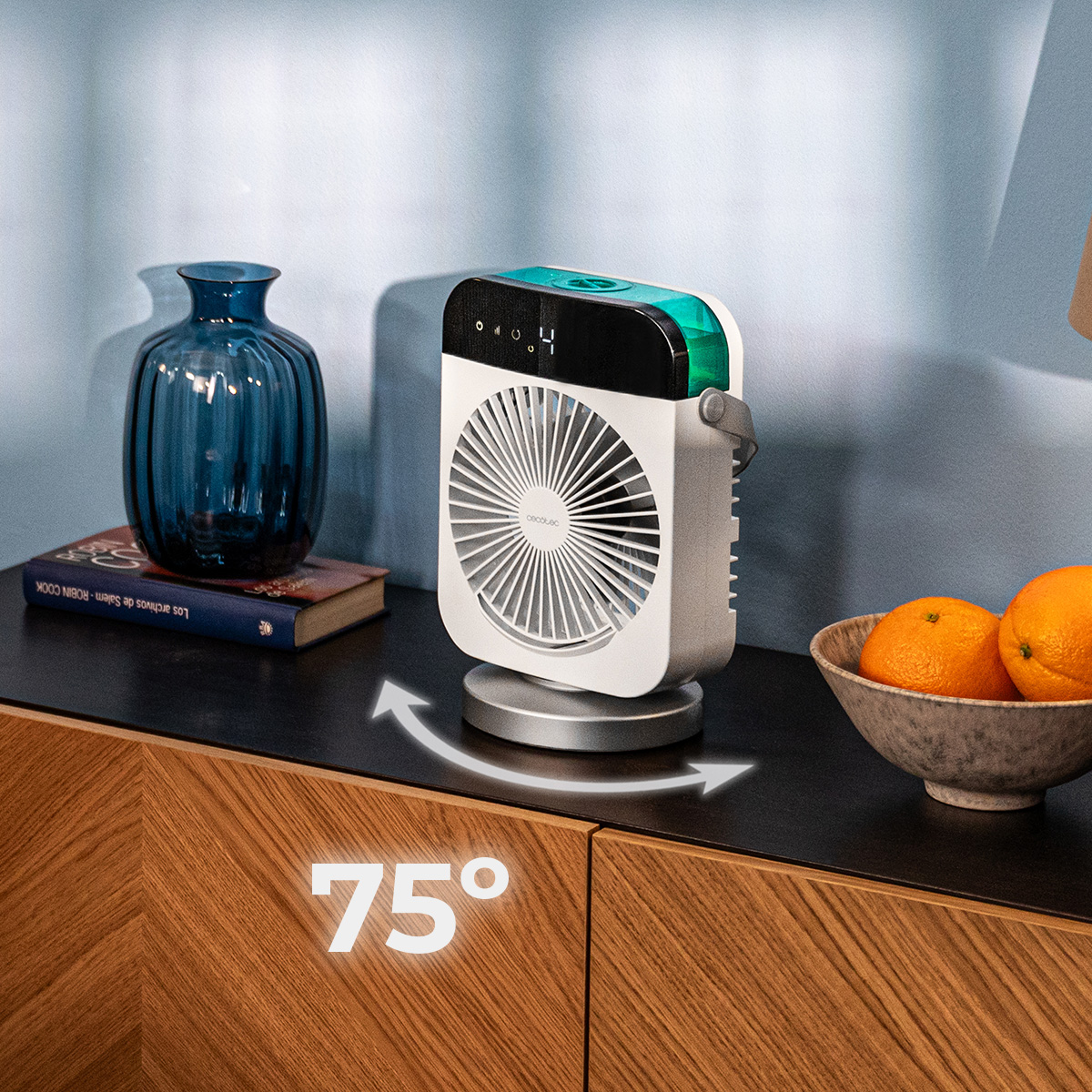 Un ventilador portátil blanco con un icono de rotación de 75 grados está sobre un mueble oscuro junto a un florero azul y naranjas.