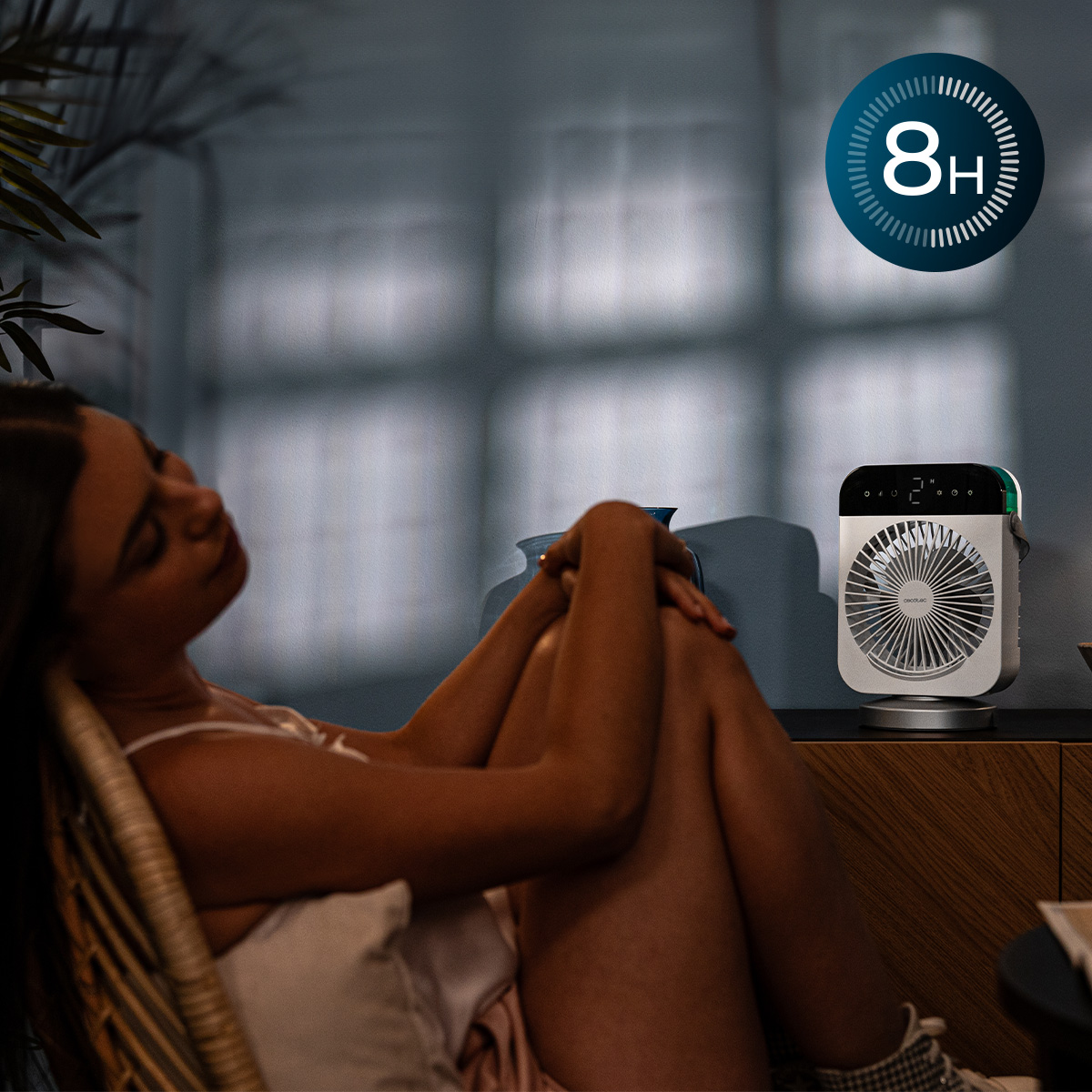 Una mujer descansa en una silla junto a una mesa pequeña con un ventilador de escritorio en una habitación oscura.