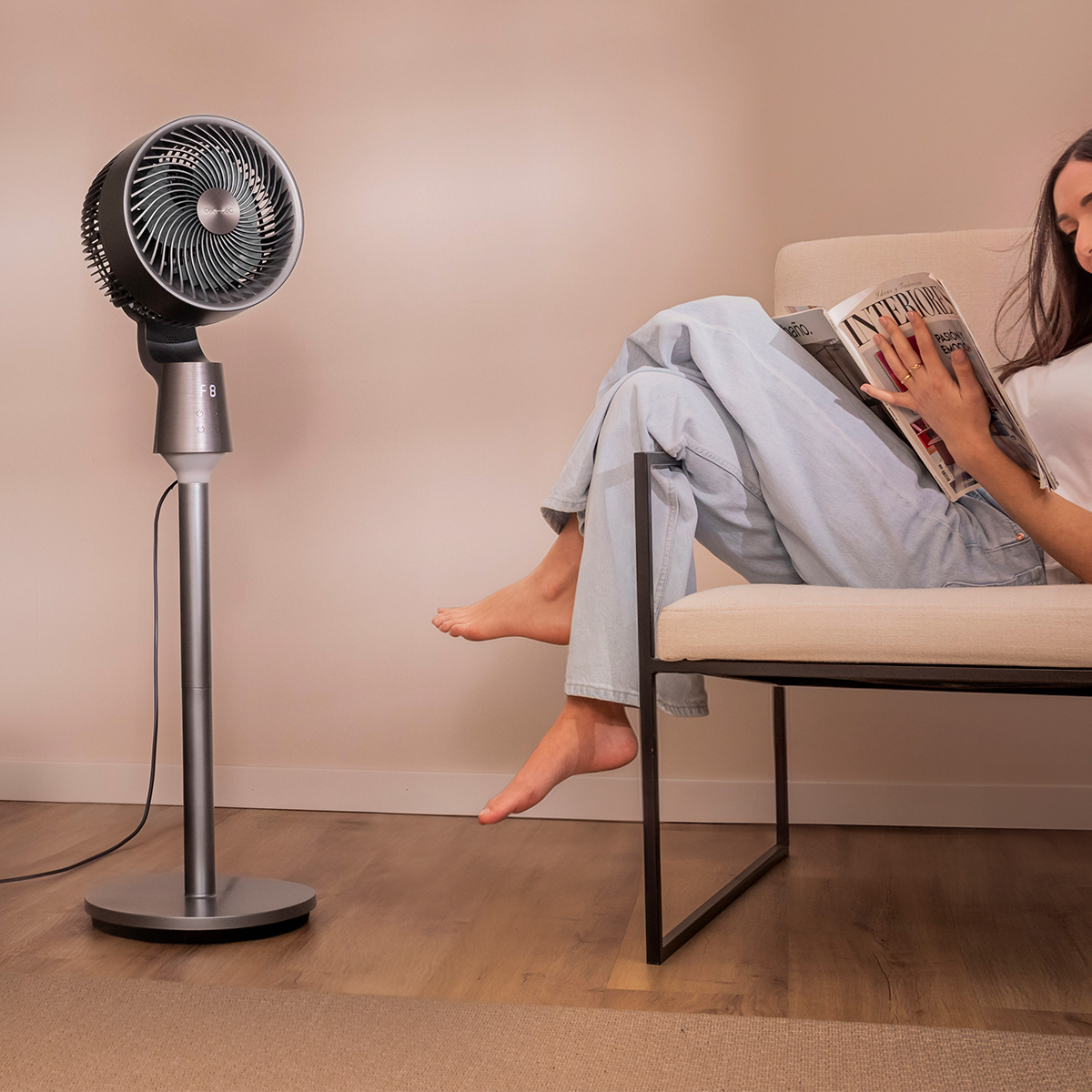 Mujer en sillón claro leyendo una revista junto a un ventilador de pie.