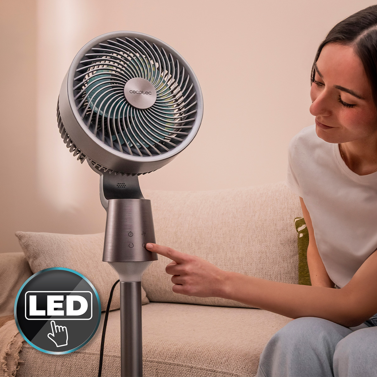 Una mujer sentada en un sofá toca el botón de encendido de un ventilador de pie gris moderno.