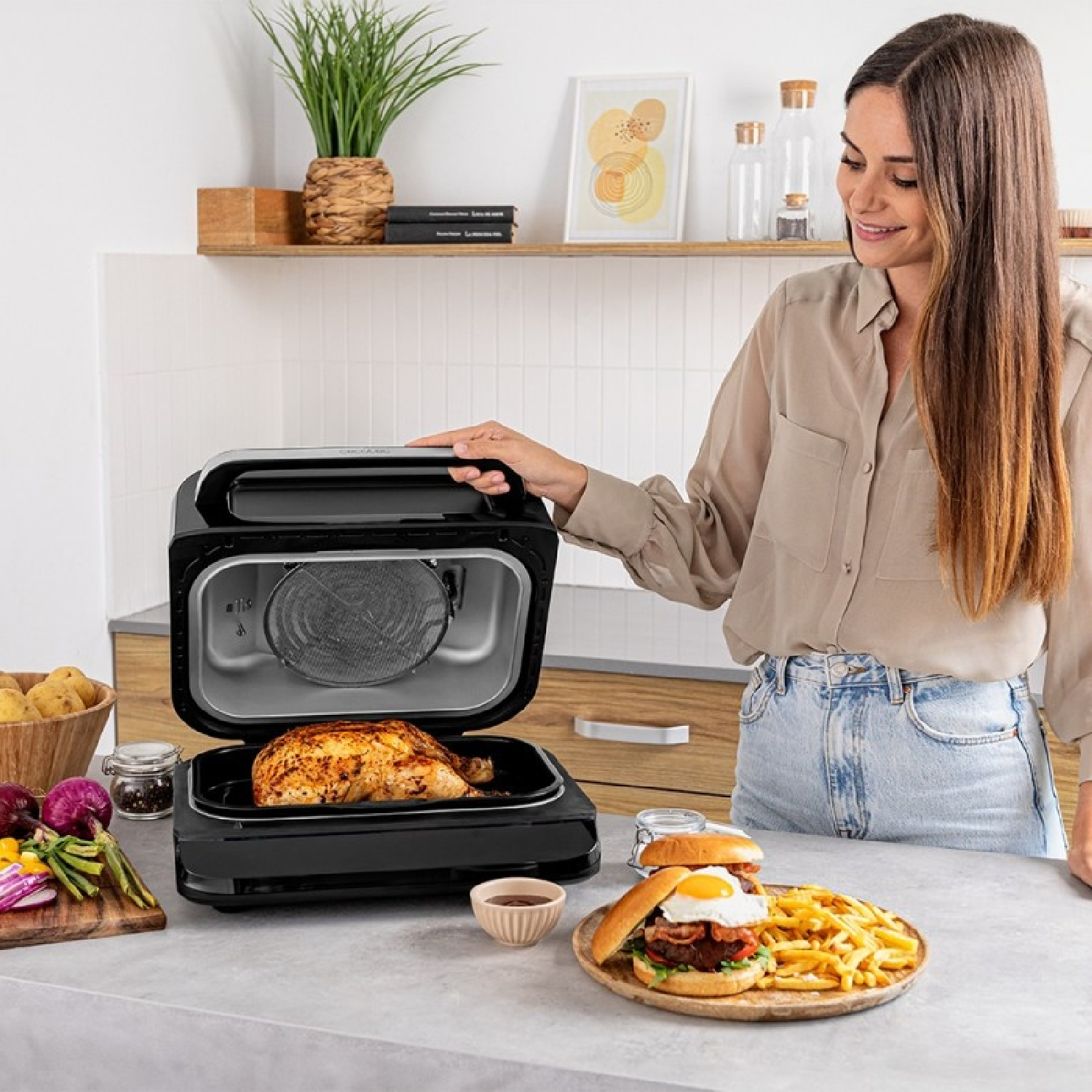Frau öffnet eine Heißluftfritteuse mit Hähnchen. Davor stehen Burger und Pommes.