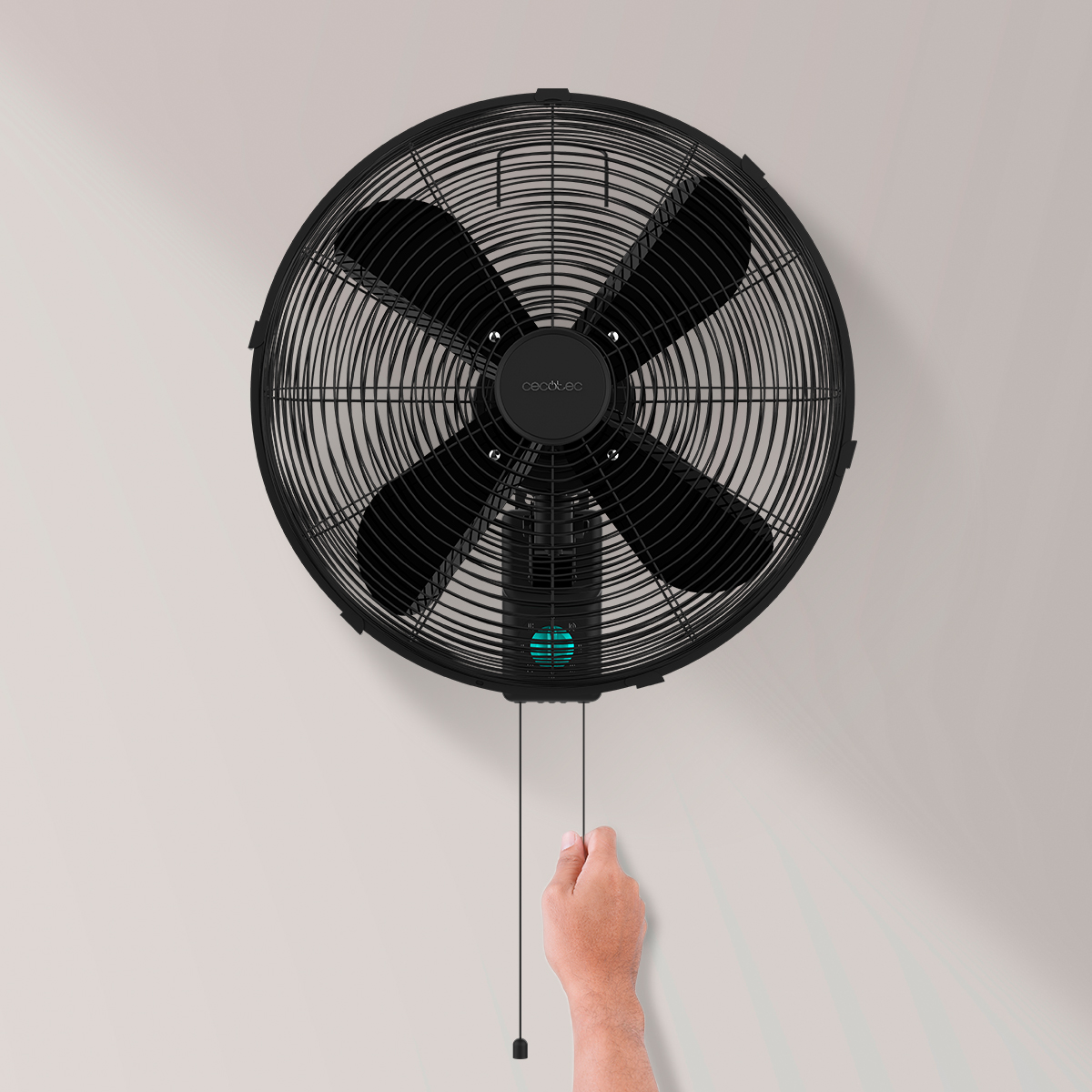 Una mano tira del cordón de un ventilador de pared negro sobre fondo liso.