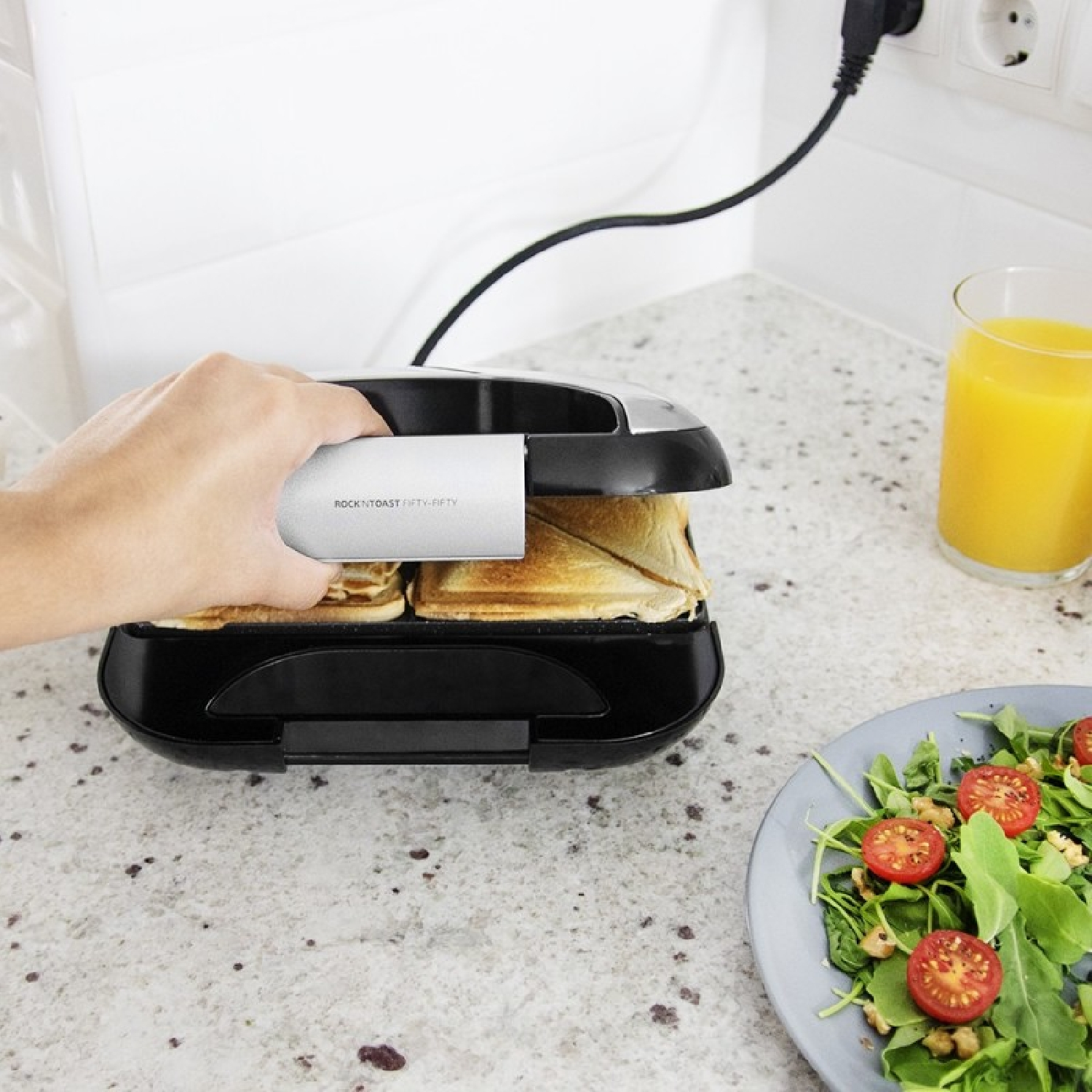 Una mano apre una piastra per panini nera e argento su un piano cucina con insalata e succo.