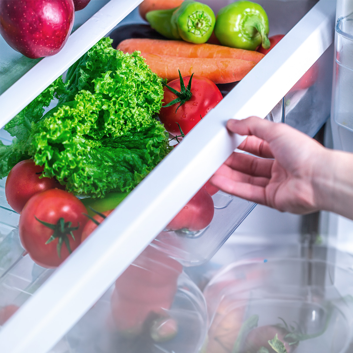 Una mano abre un cajón de refrigerador lleno de frutas y verduras frescas.