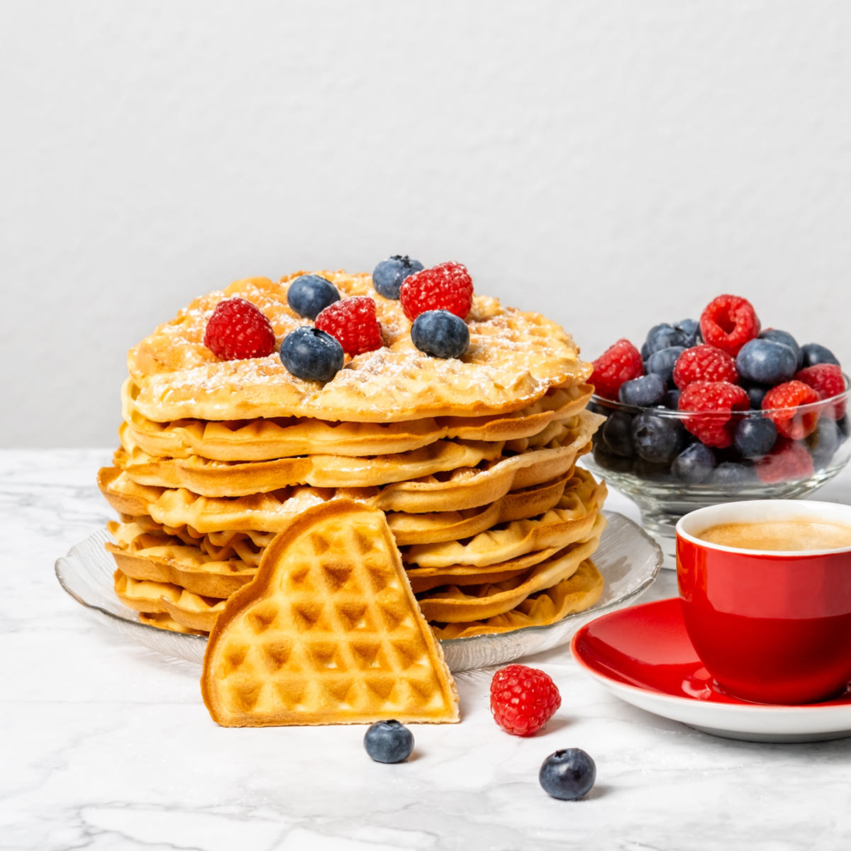 Pila di waffle dorati con bacche fresche, zucchero a velo, un waffle a forma di cuore separato e tazza di caffè rossa.