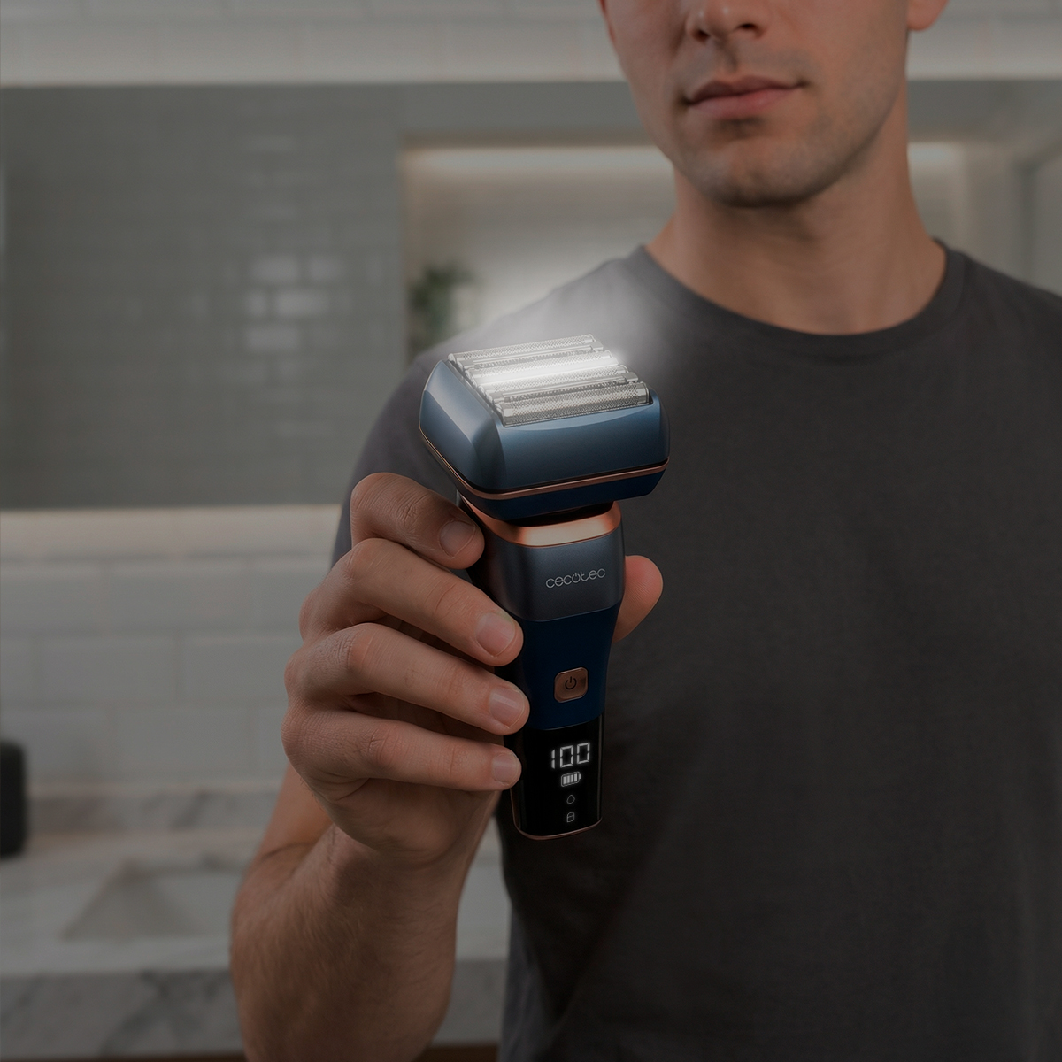 Un hombre con una camiseta gris sostiene una afeitadora eléctrica azul y cobre con pantalla digital.