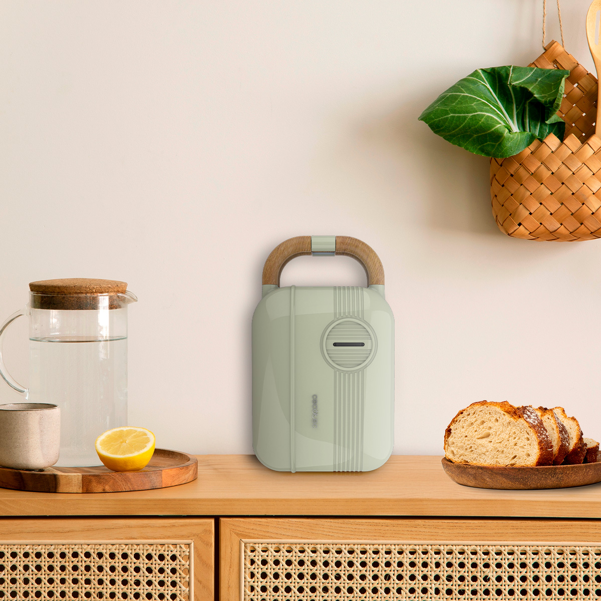 Una tostiera verde pastello su un bancone di legno con caraffa d'acqua, limone, pane e cestino sospeso.