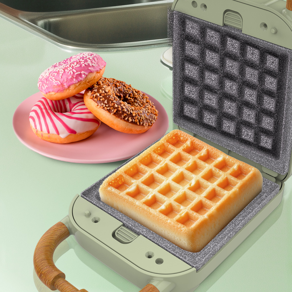 Una piastra per waffle con un waffle appena cotto all'interno, accanto a un piatto rosa con tre ciambelle glassate su un bancone verde.