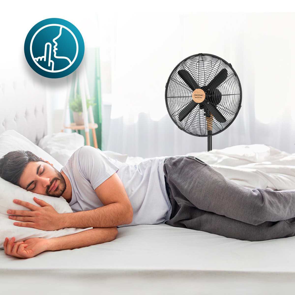 Un hombre duerme plácidamente en la cama con un ventilador de pie negro silencioso.