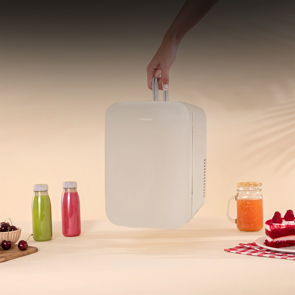 Una mano sostiene una mininevera beige sobre una mesa con jugos, cerezas y pastel.