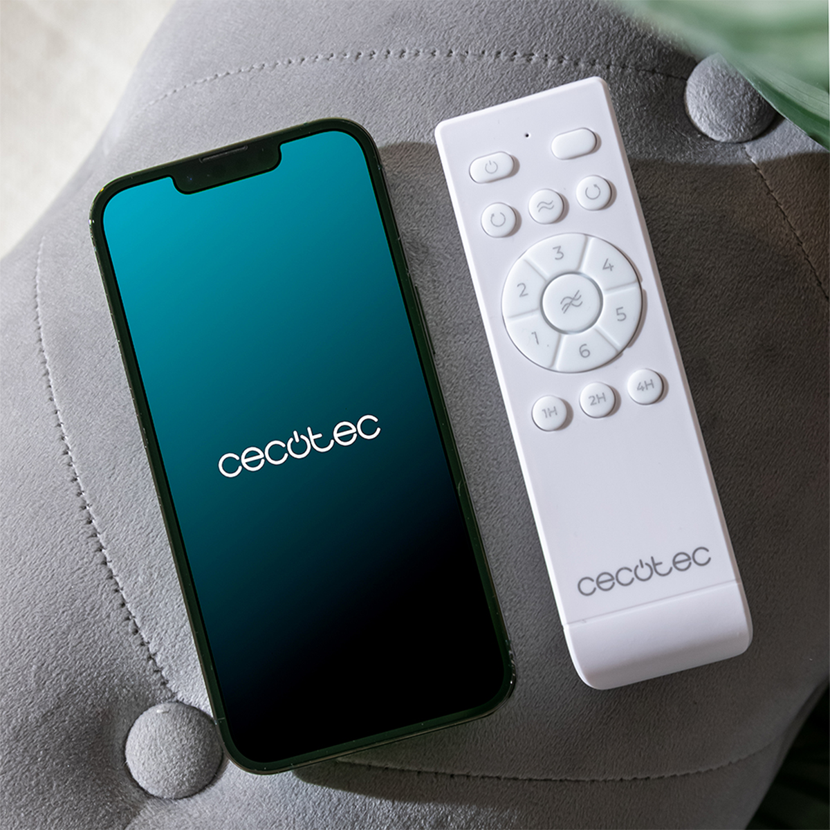Uno smartphone nero con logo Cecotec e un telecomando bianco Cecotec su un divano grigio.