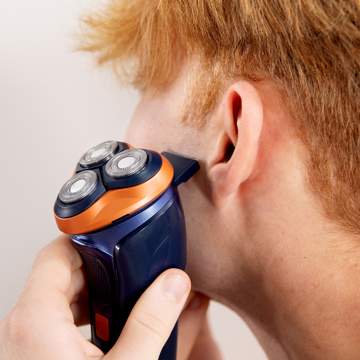 Un hombre pelirrojo se afeita las patillas con una afeitadora eléctrica azul y naranja.