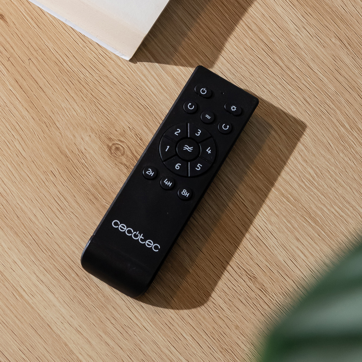 Un telecomando nero Cecotec con pulsanti rotondi e funzioni timer poggia su una superficie in legno chiaro.
