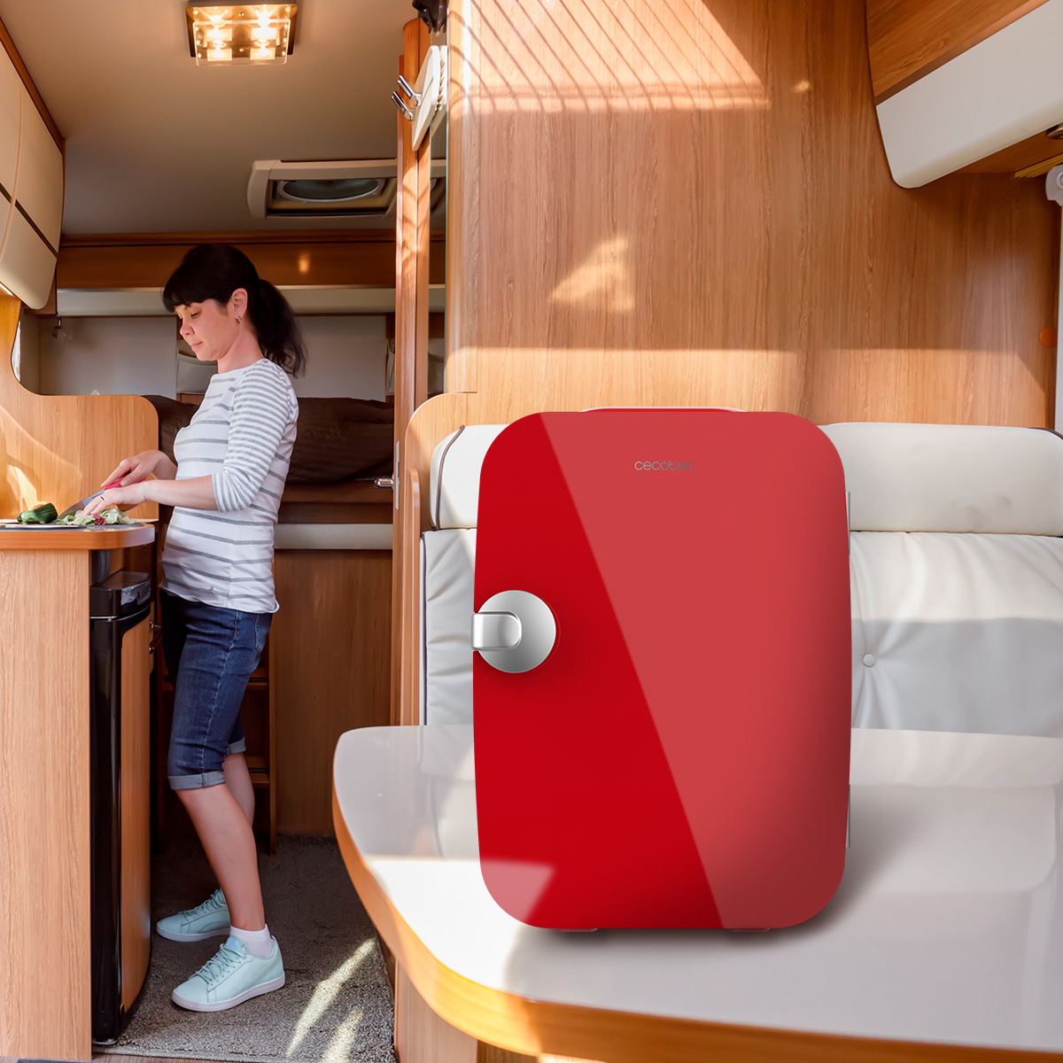 Una mini nevera roja en una mesa blanca dentro de una autocaravana, con una mujer preparando comida.