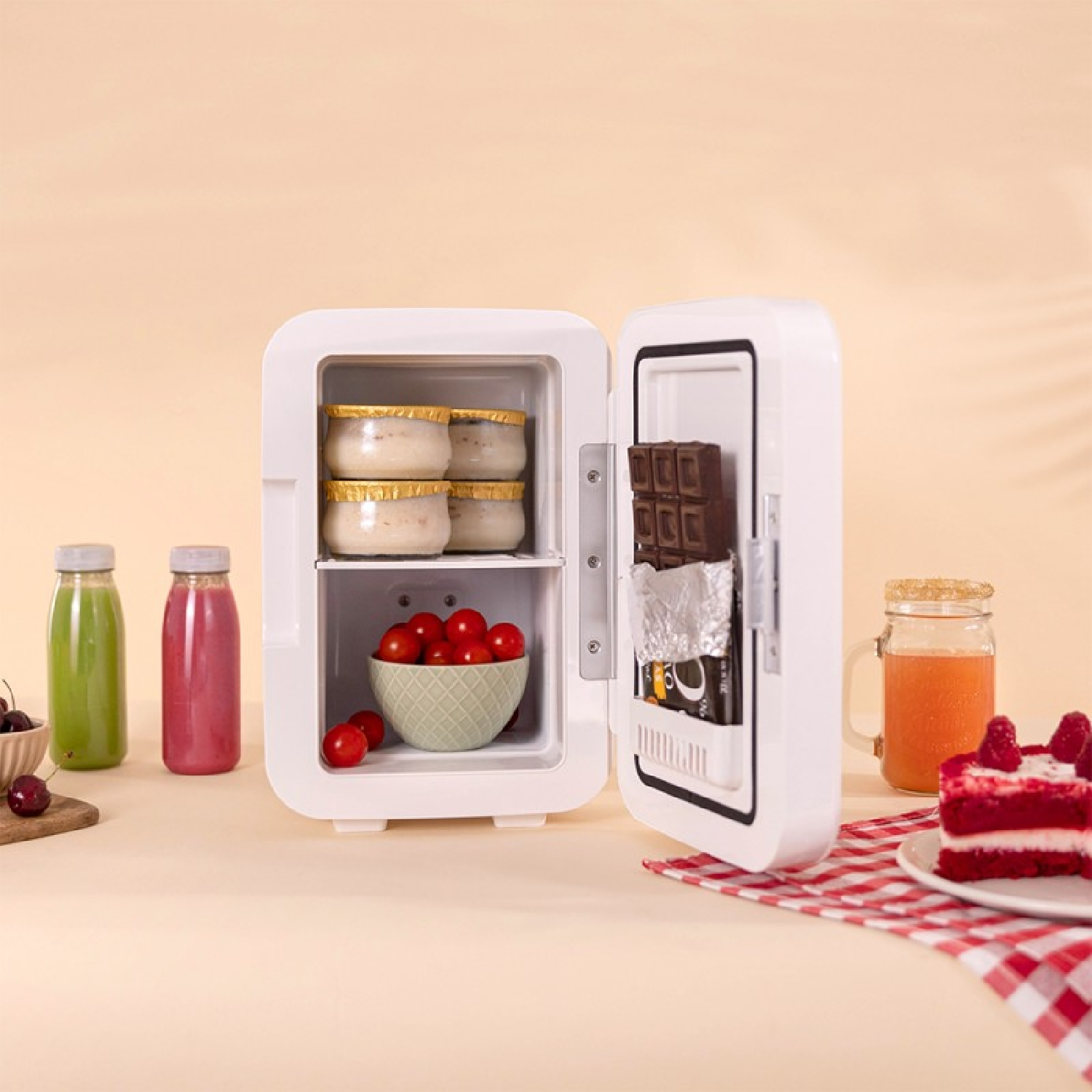 Piccolo mini frigo bianco aperto con yogurt, pomodori e cioccolato, circondato da bevande e torta.