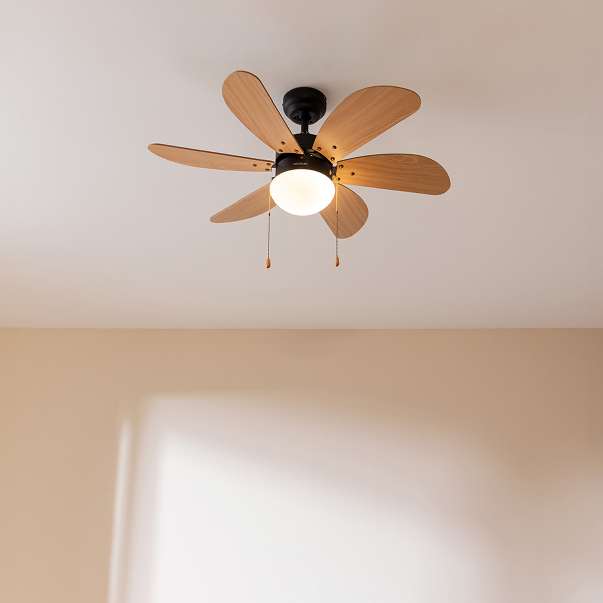 Un ventilatore da soffitto bianco con sei pale in legno e una luce illuminata al centro pende da un soffitto bianco.