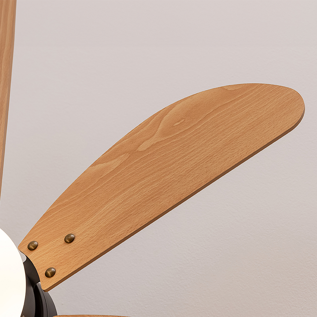 Primo piano di una pala di ventilatore a soffitto in legno con viti in ottone contro una parete bianca.