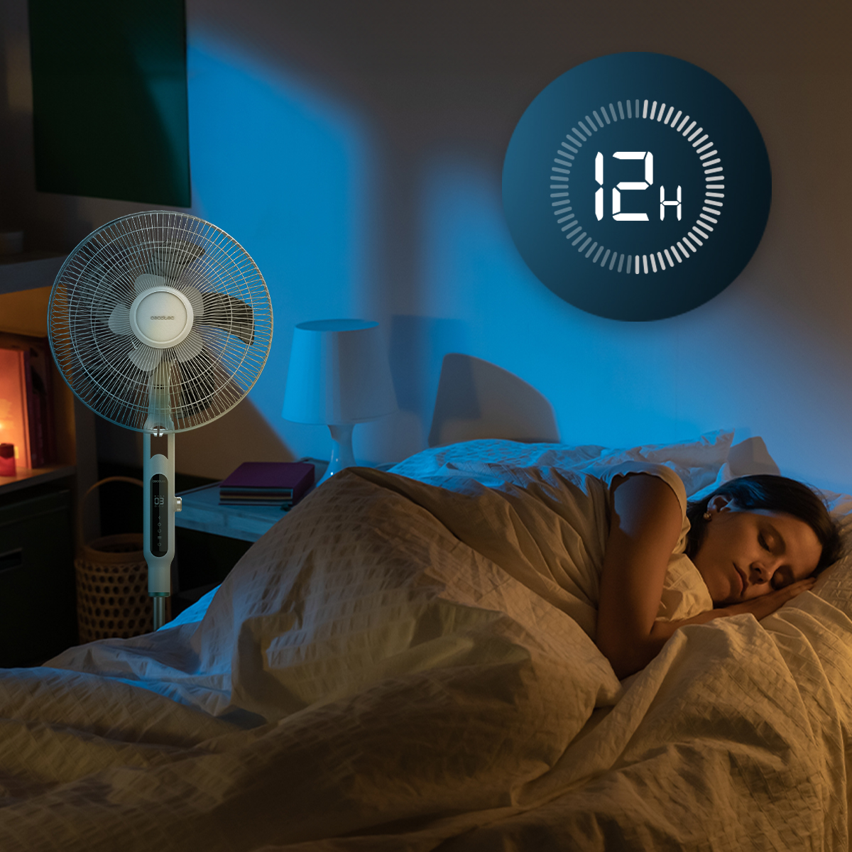 Una mujer duerme en la cama mientras un ventilador de pie funciona cerca en una habitación tenuemente iluminada.