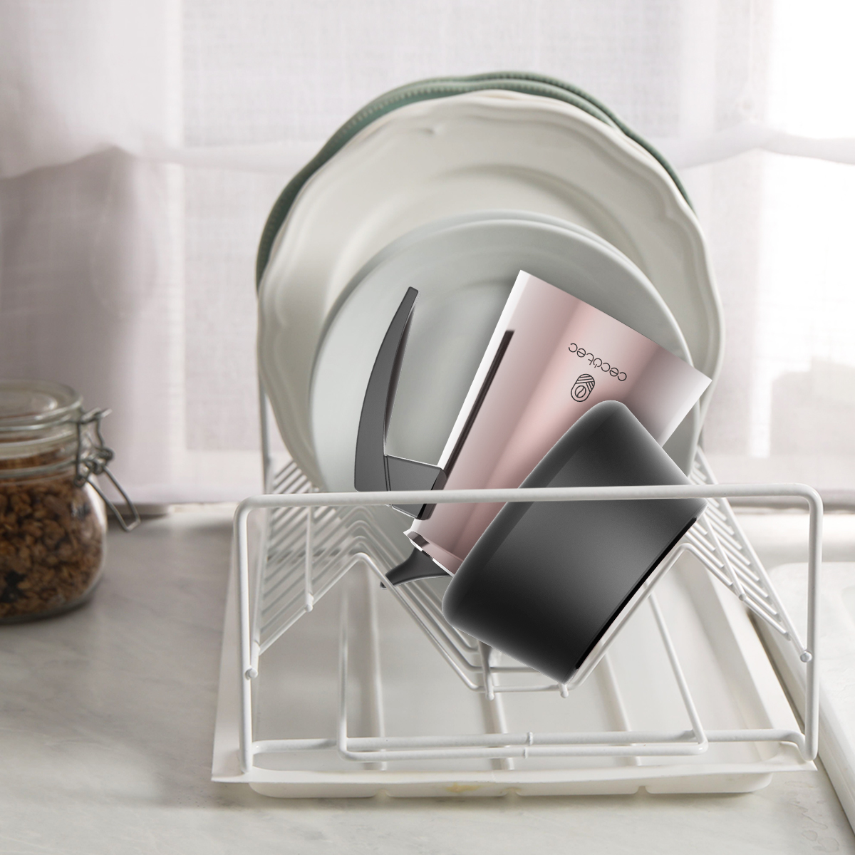 Una caffettiera rosa e una tazza nera in uno scolapiatti bianco con piatti dietro su un bancone.