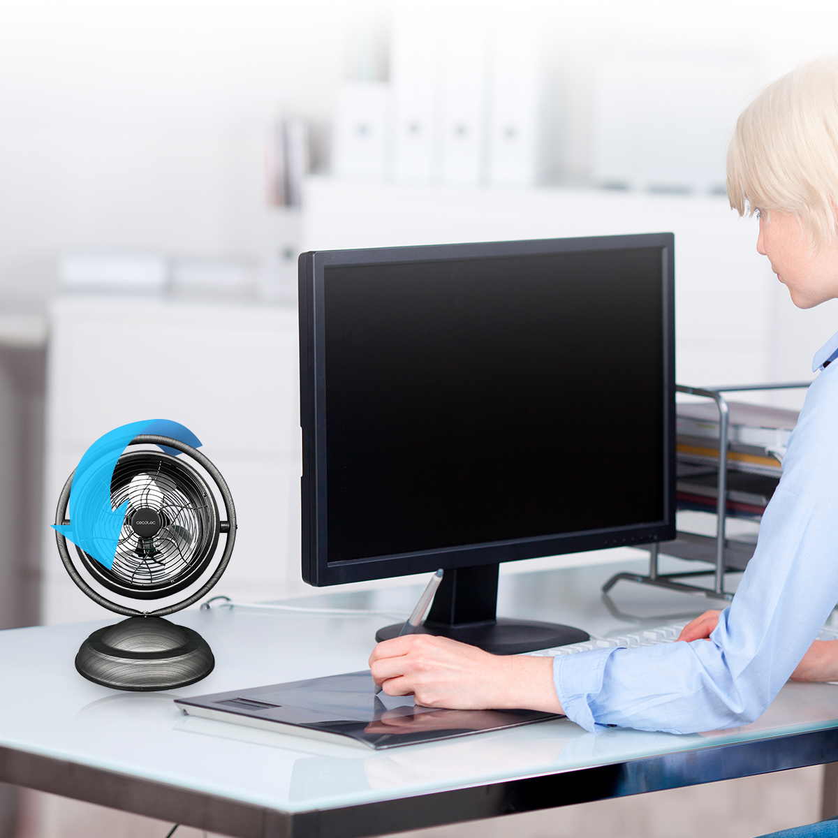 Una donna lavora a una scrivania con computer, tavoletta grafica e un piccolo ventilatore.