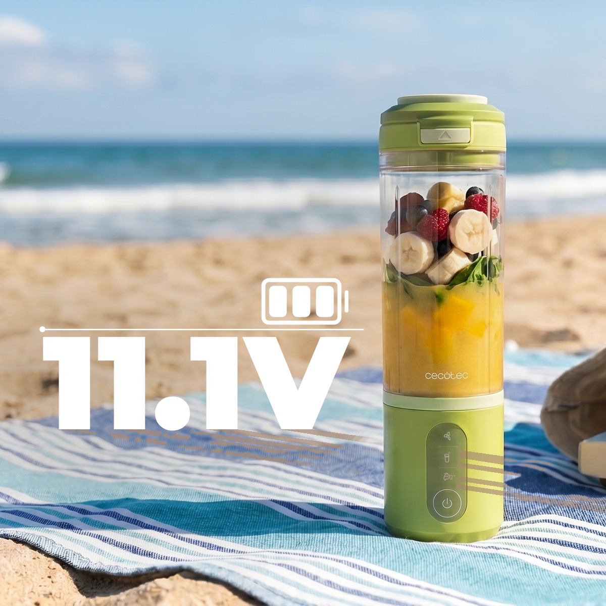 Licuadora portátil verde con fruta y jugo sobre una toalla de playa, con el mar de fondo.