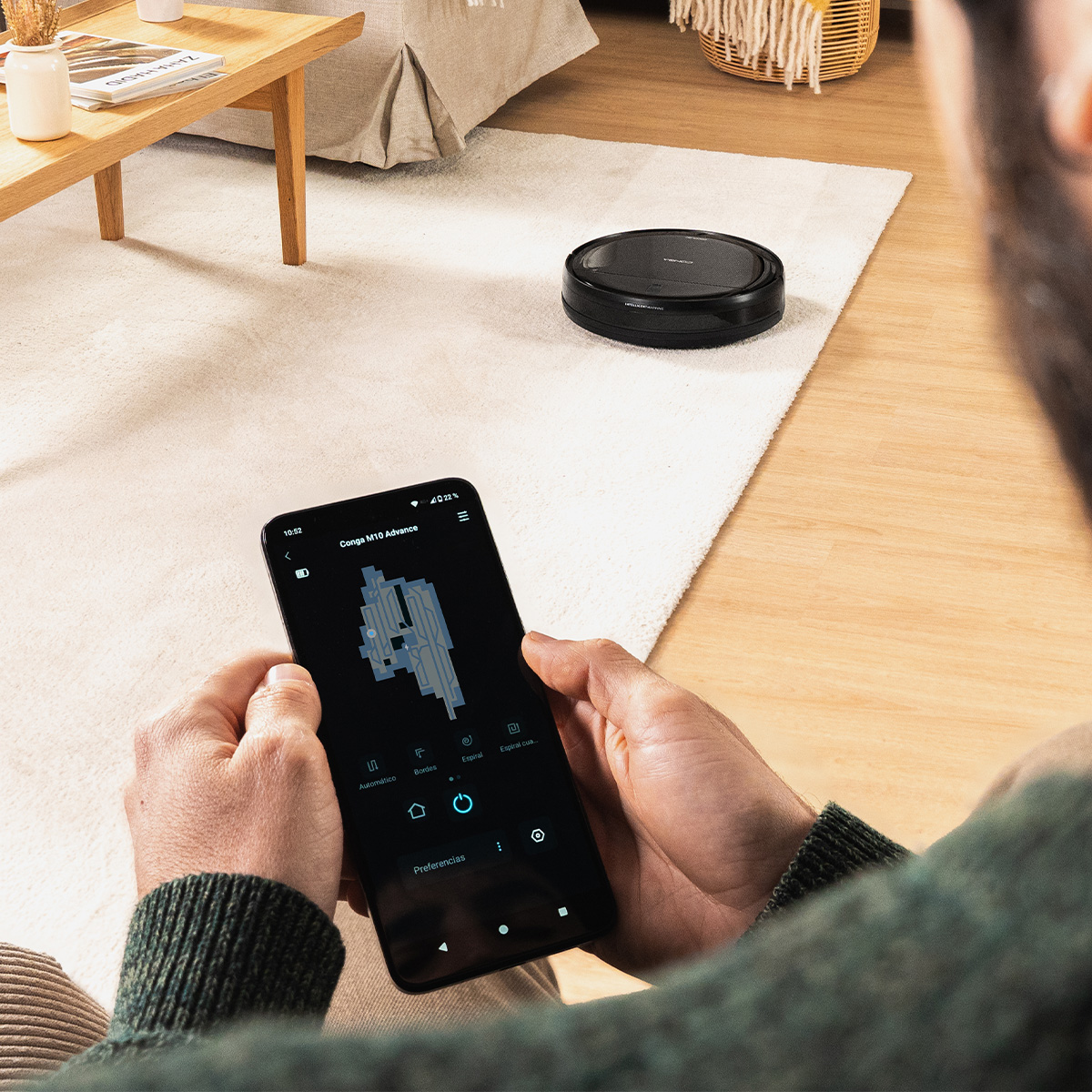 Un uomo usa un'app sullo smartphone per controllare un robot aspirapolvere nero su un tappeto chiaro.