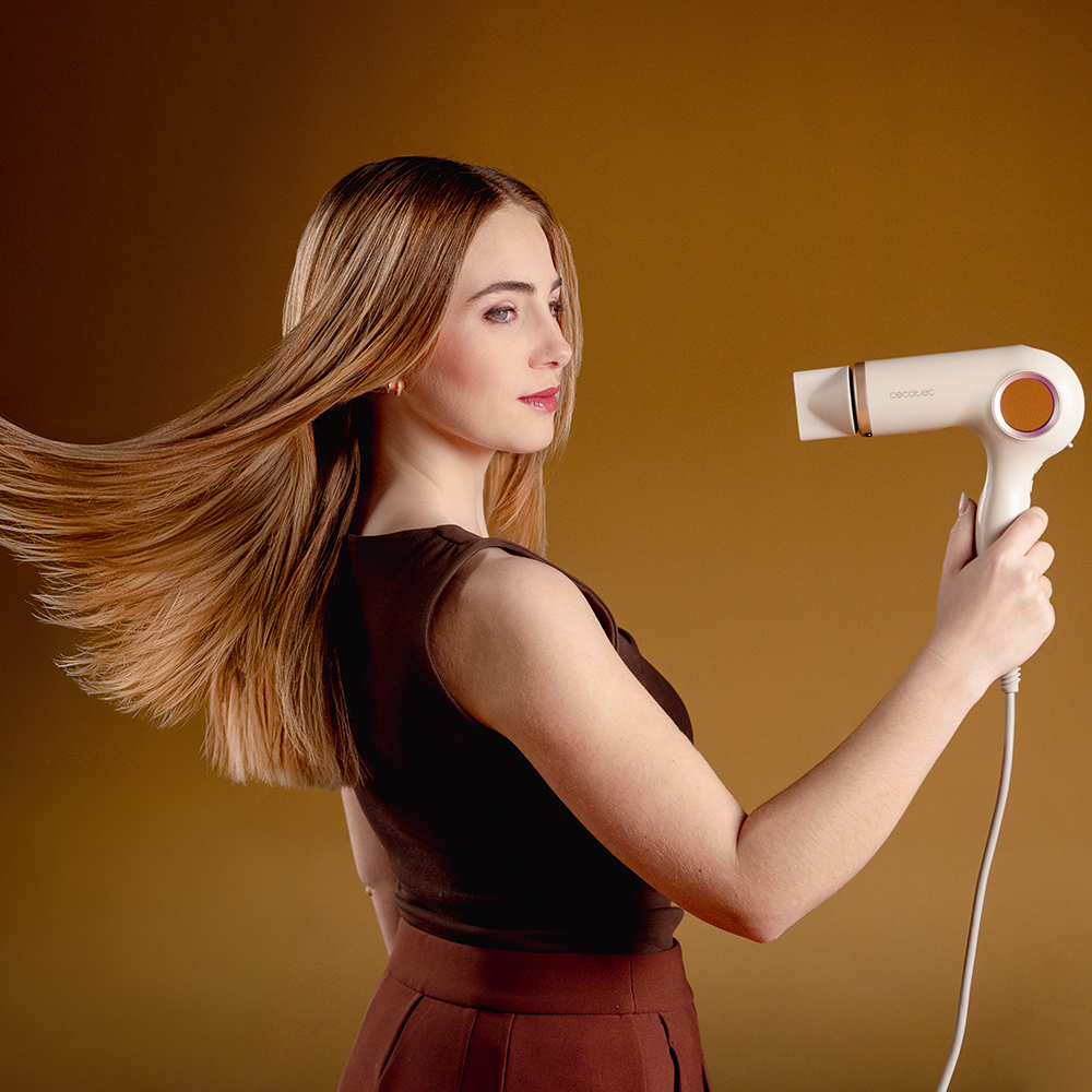 Una mujer con top marrón y pelo largo sostiene un secador de pelo blanco moderno sobre fondo ocre.
