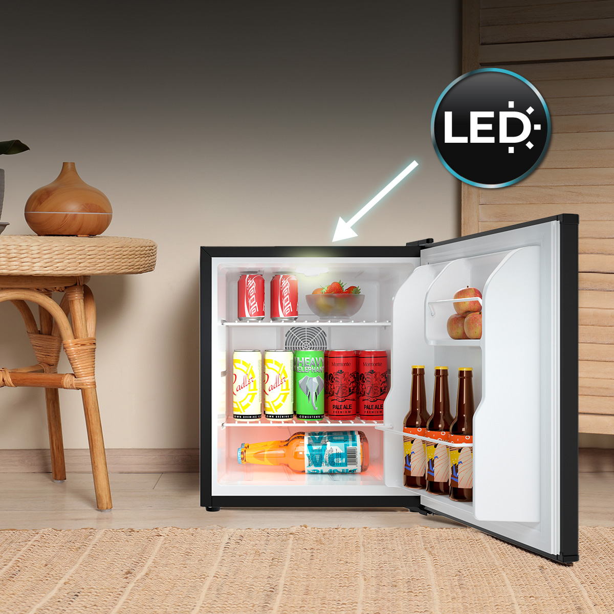 Pequeño mininevera abierta en suelo de madera con latas, botellas y frutas, iluminada por una luz LED.