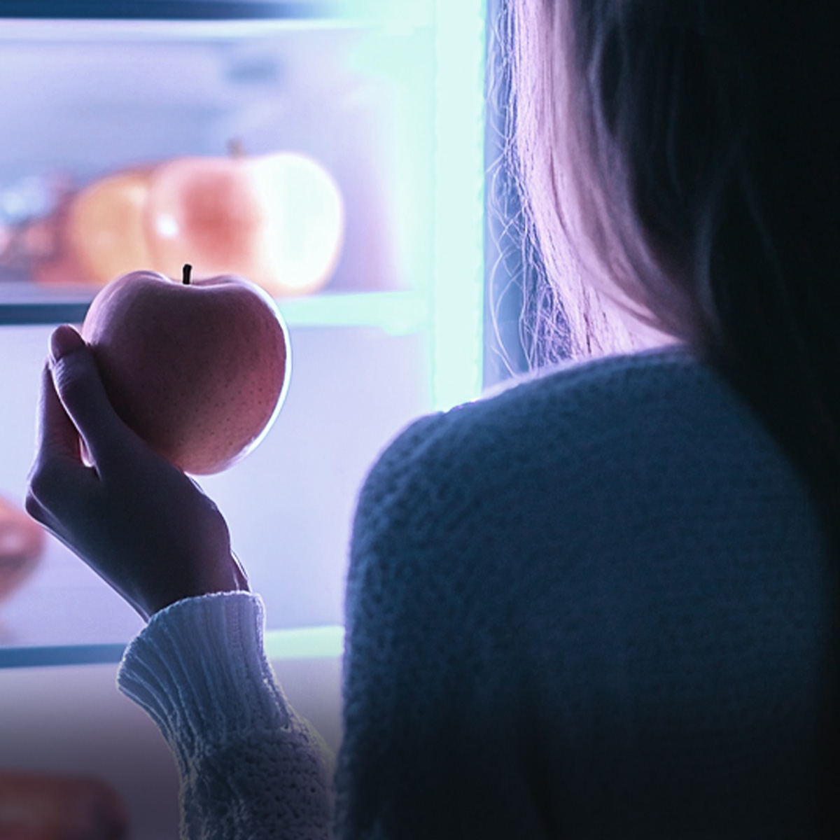 Una persona sostiene una manzana roja frente a un refrigerador abierto e iluminado por la noche.