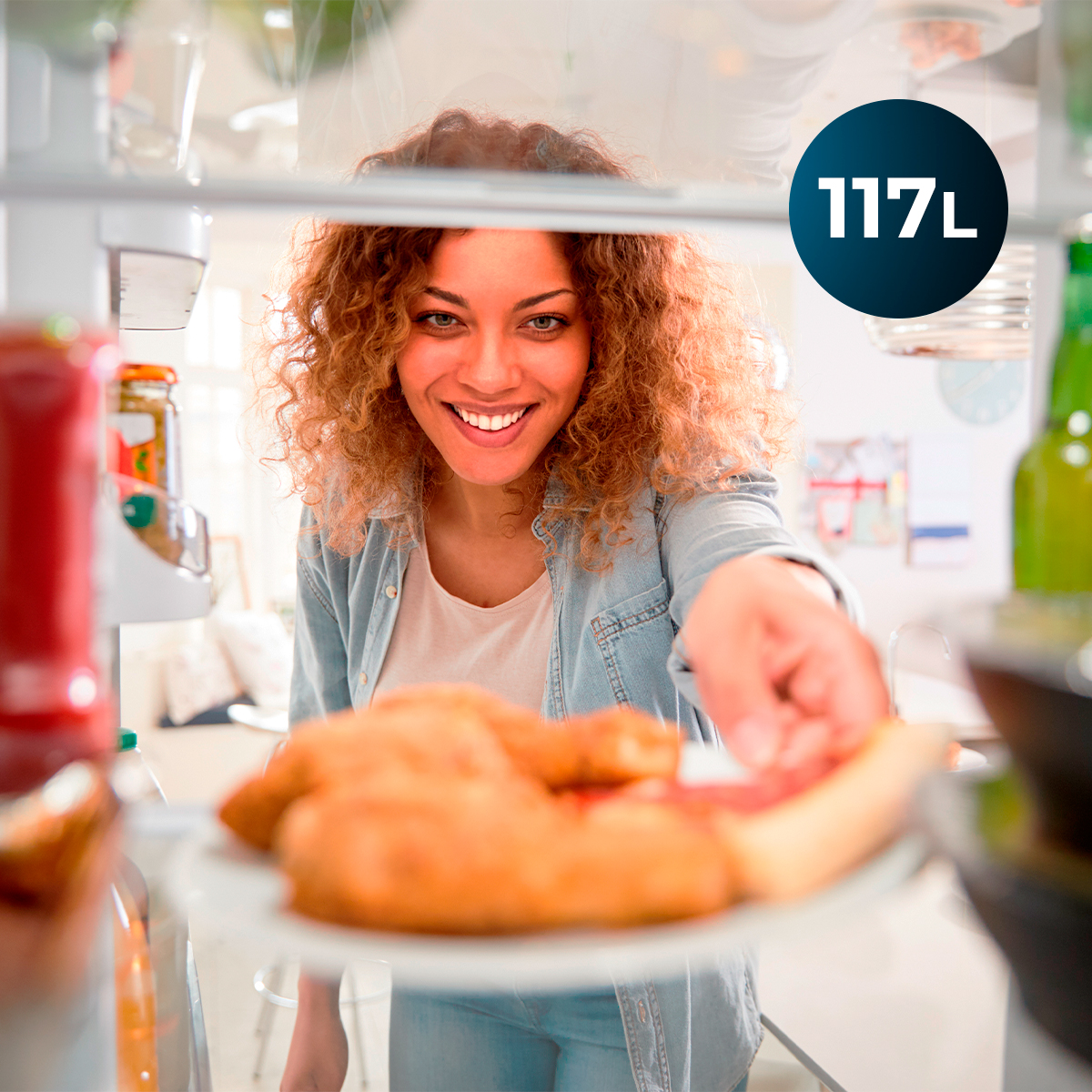 Mujer sonriente abre un refrigerador y sostiene un plato con pollo horneado.