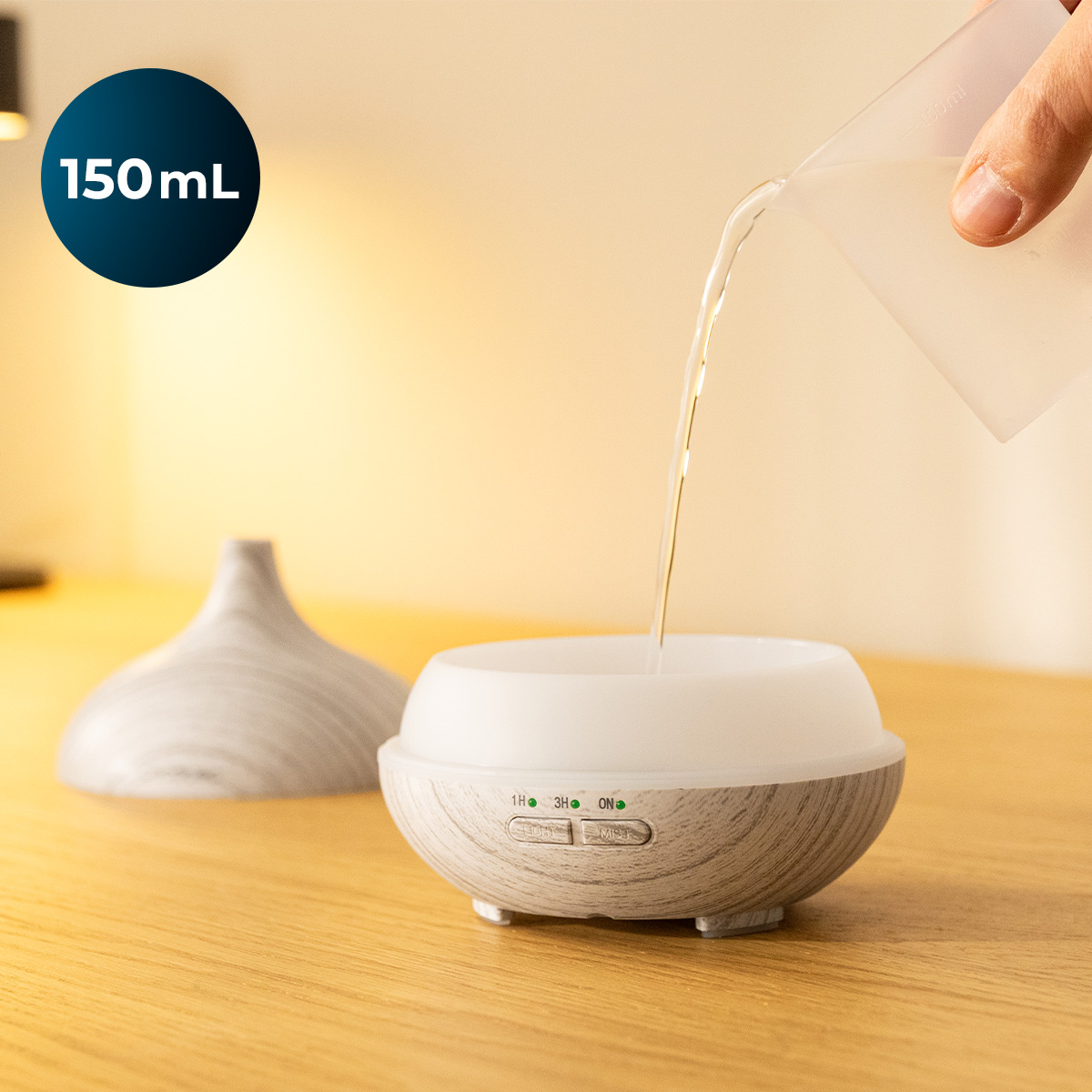 Wasser wird aus einem Messbecher in den offenen Behälter eines Aroma-Diffusers auf einem Holztisch gegossen.