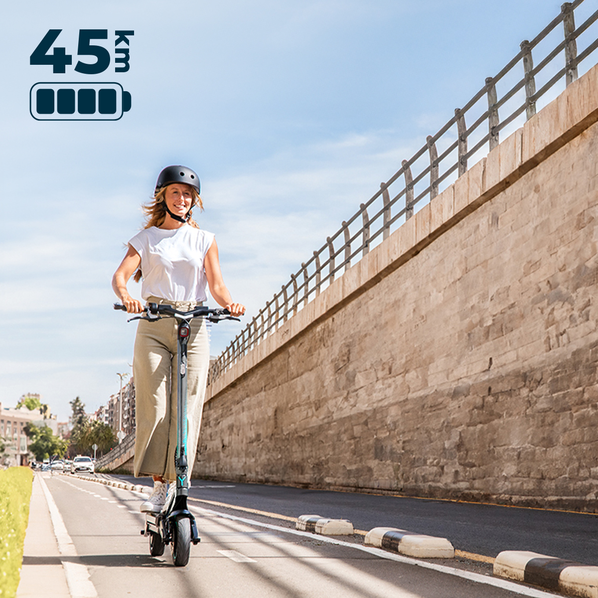 Una donna sorridente con casco guida un monopattino elettrico su una pista ciclabile vicino a un muro.