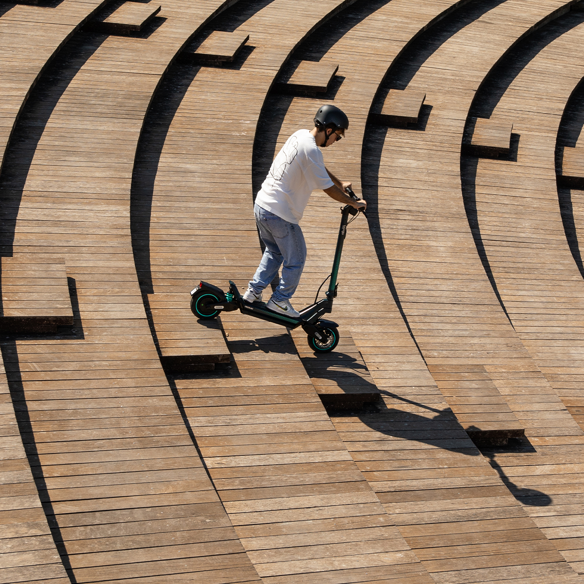 Una persona con casco conduce un patinete eléctrico por un gran anfiteatro de madera escalonado.