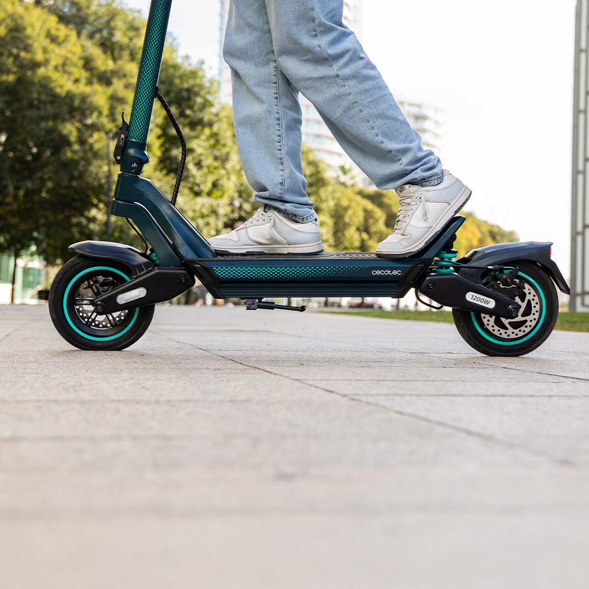 Persona con jeans claros y zapatillas montando un patinete eléctrico verde oscuro en un camino pavimentado.