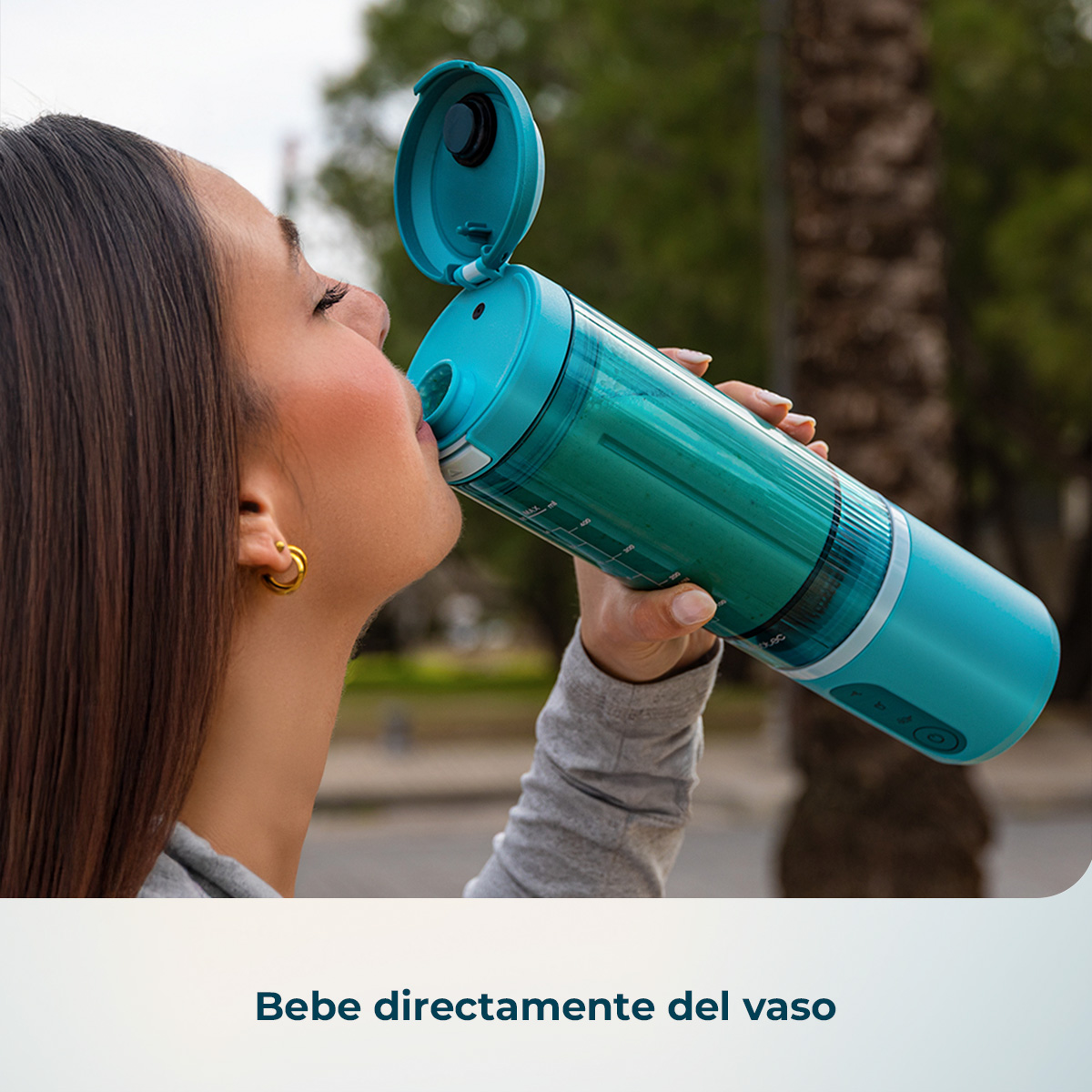 Una mujer con cabello castaño largo bebe de una botella de licuadora portátil azul al aire libre.
