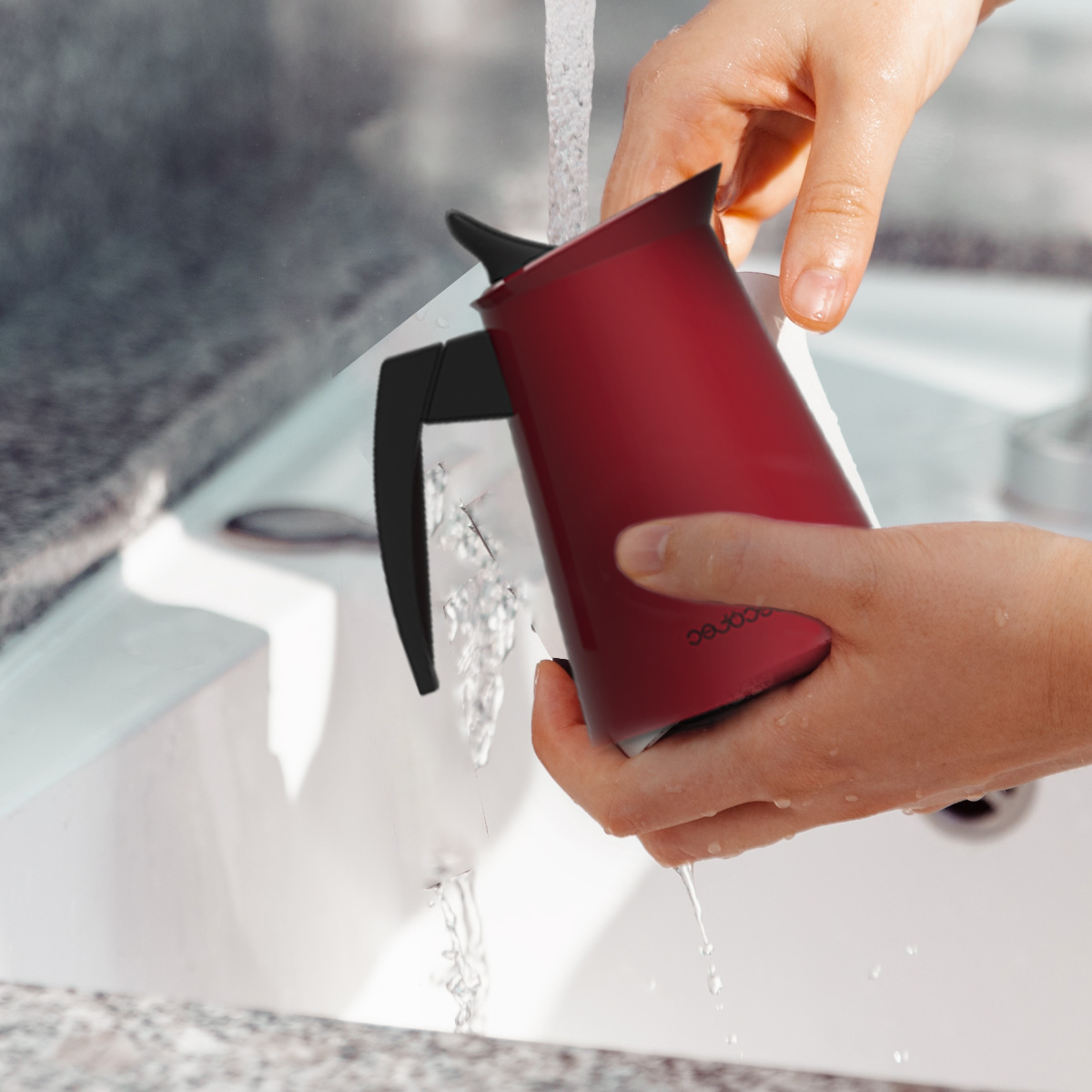 Una persona lavando una cafetera roja con asa negra bajo el agua corriente en un lavabo.