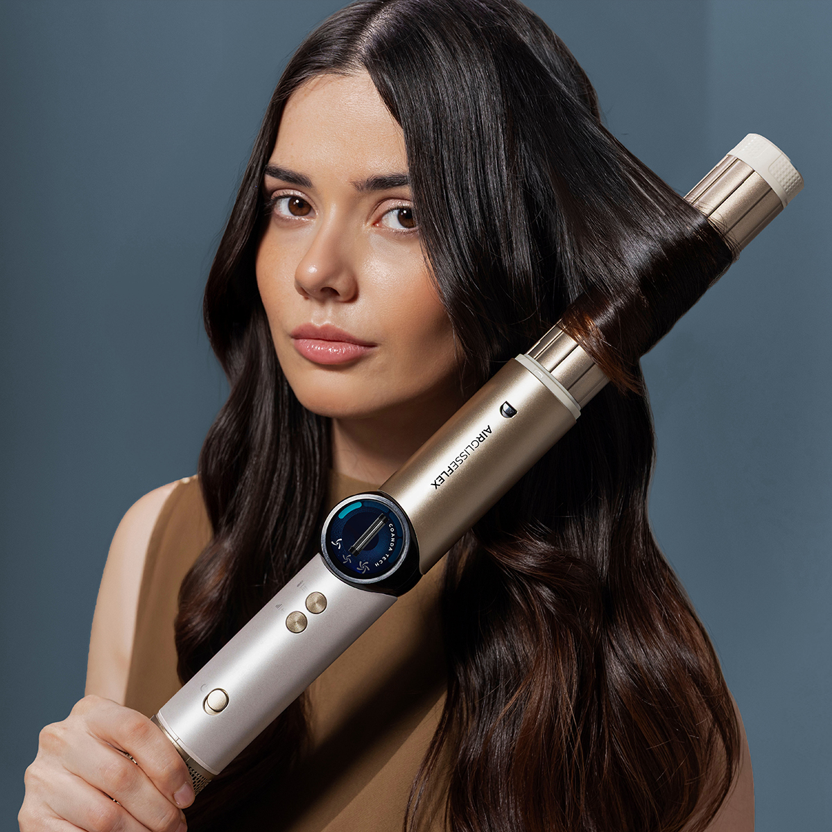 Una mujer con cabello largo y oscuro lo peina con un rizador dorado sobre fondo azul.