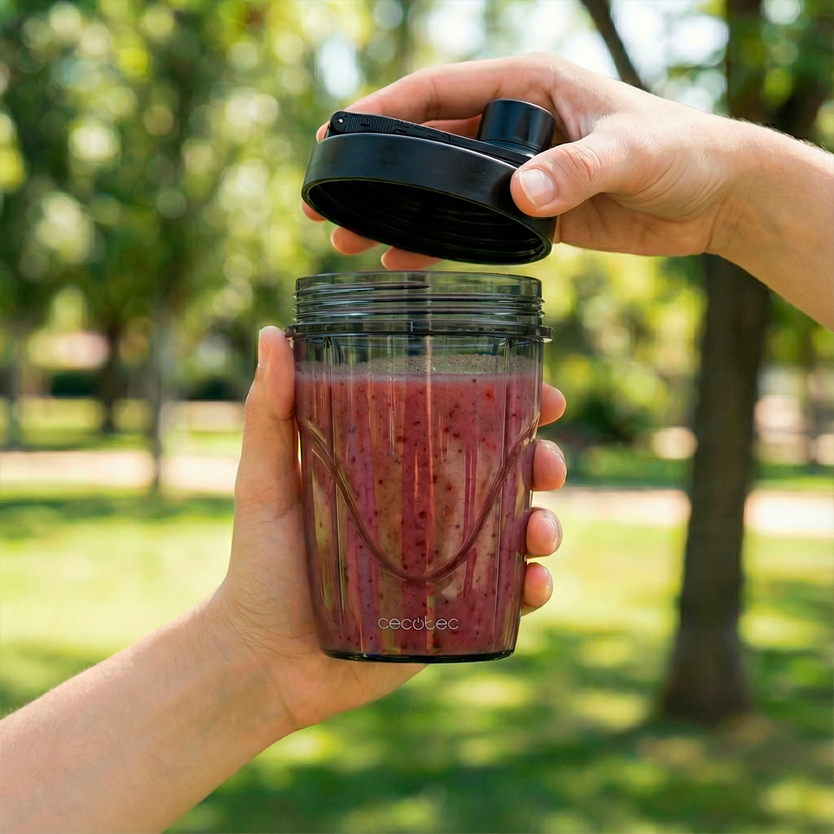 Persona abriendo un vaso mezclador con batido rojo al aire libre con fondo natural.