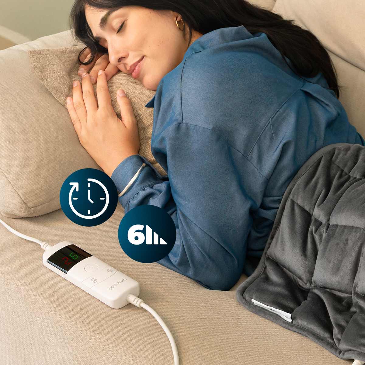 Una donna dorme su un divano, coperta da un cuscino riscaldato grigio collegato a un telecomando.