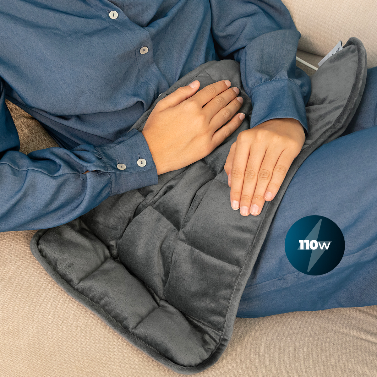 Persona con camicia blu su un divano tiene un termoforo grigio da 110W.