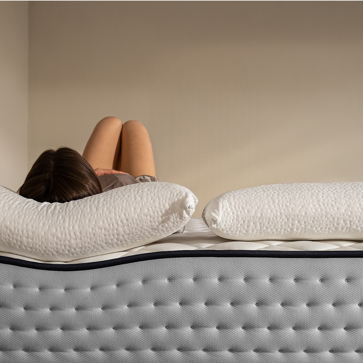 Una mujer yace en la cama sobre almohadas blancas en un colchón gris.