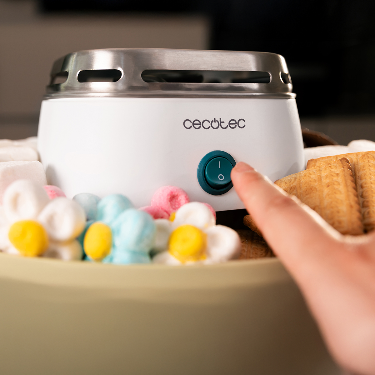 Un dito preme il pulsante di accensione di una fontana di cioccolata bianca Cecotec, circondata da caramelle e biscotti.