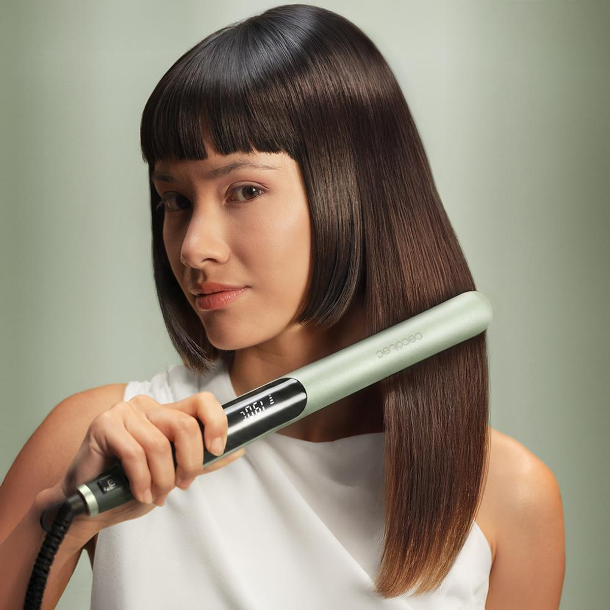 Una donna con capelli castani lisci tiene una piastra per capelli verde, guardando la fotocamera.